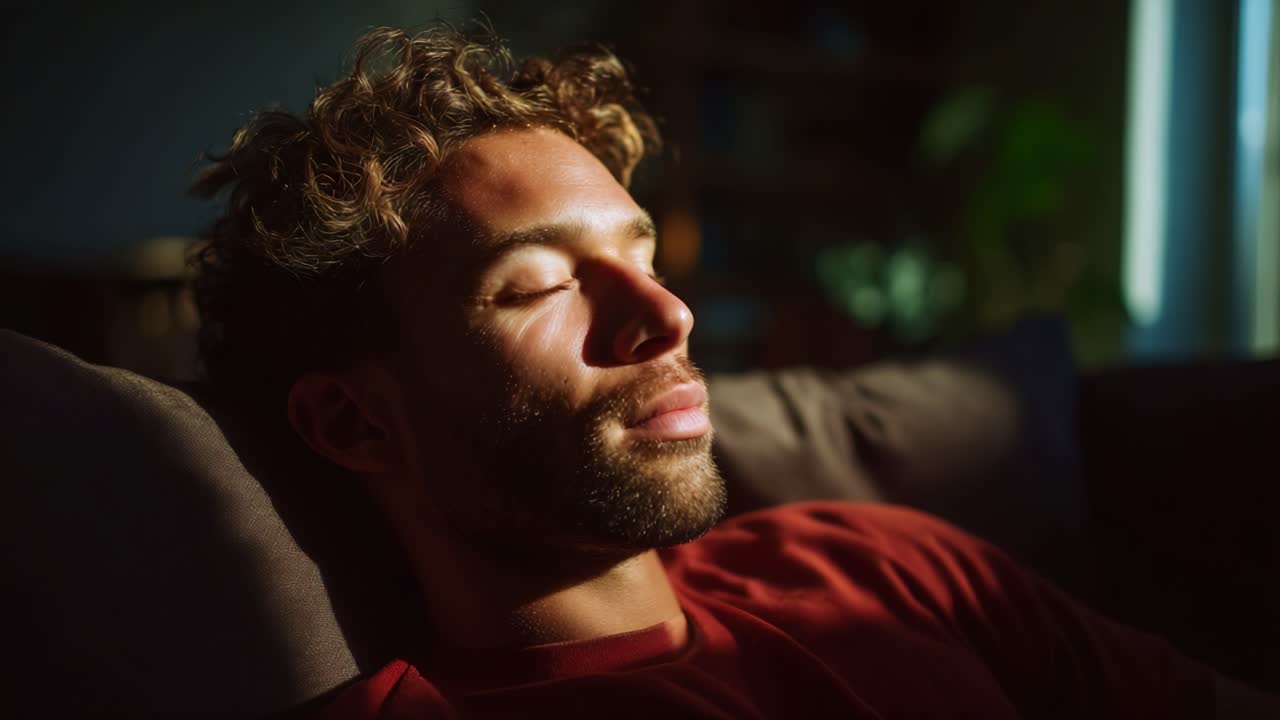 En avslappet mann nyter et øyeblikk av fredelig ro mens han sitter på en sofa, badet i mykt, naturlig lys, noe som skaper en drømmeaktig og rolig atmosfære