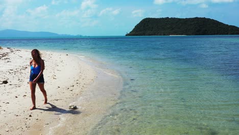 Attractive-woman-walking-alone-on-white-sandy-beach-on-a-windy-morning-alongside-calm-clear-water-of-blue-turquoise-lagoon