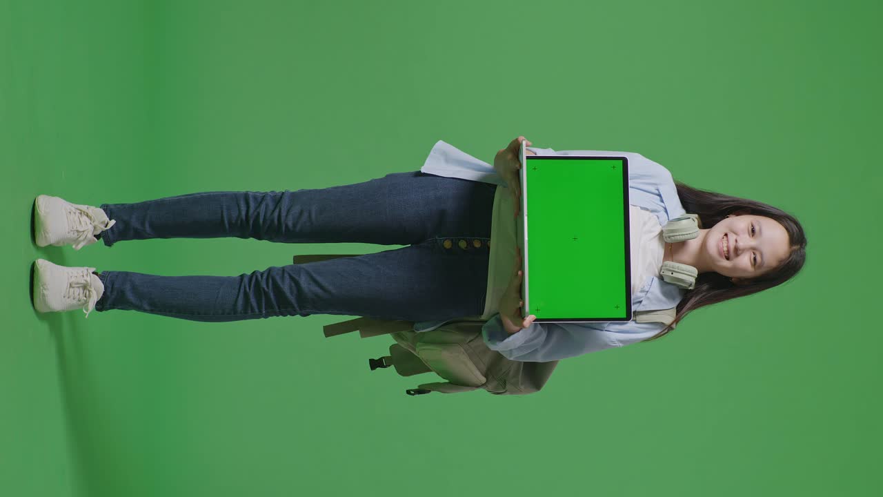cuerpo lleno de estudiante adolescente asiática con una mochila sonriendo y mostrando portátil de pantalla verde a la cámara mientras está de pie en el estudio de fondo de pantalla verde