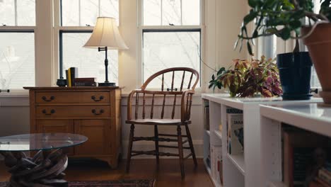 a-wooden-chair-next-to-a-wooden-chest-with-a-small-lamp-next-to-white-bookshelves-inside-of-a-sun-room-with-windows-letting-in-natural-sunlight