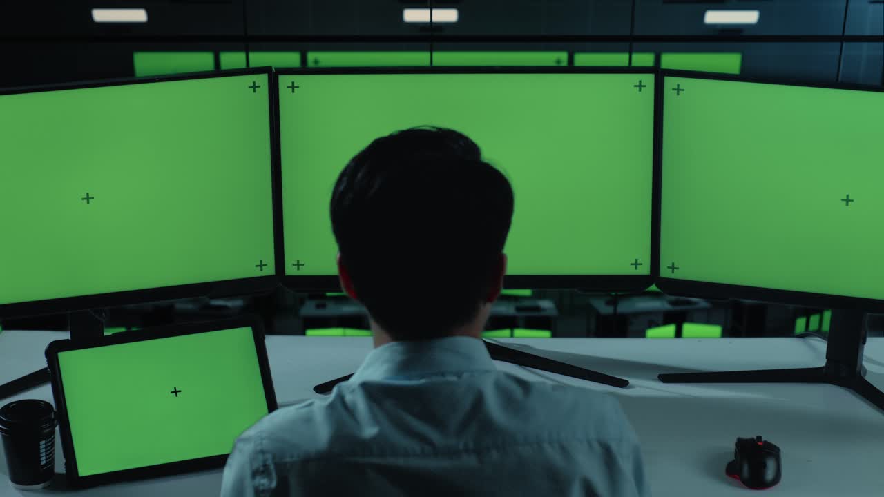 vista trasera de un hombre asiático trabajando con una tableta y varios monitores de computadora con una pantalla verde simulada en la oficina