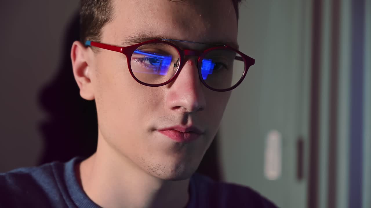 Young man in red glasses watching at computer monitor in a green and purple lighted room. Working process reflection in the lens. Warm light on half of the face