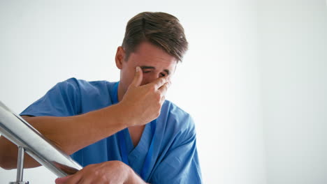 Stressed-Male-Doctor-Or-Nurse-Wearing-Scrubs-Standing-On-Stairs-In-Hospital-Suffering-With-Burnout