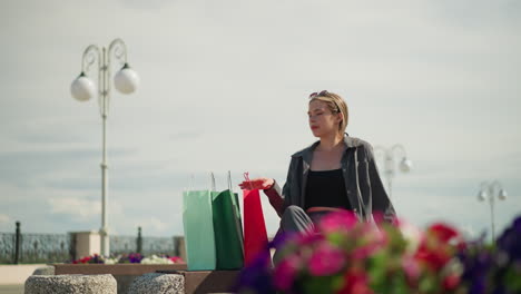 lady casually drops her shopping bags on a bench and sits down with a relaxed expression, looking off into the distance, light poles and blurred greenery are visible in the background