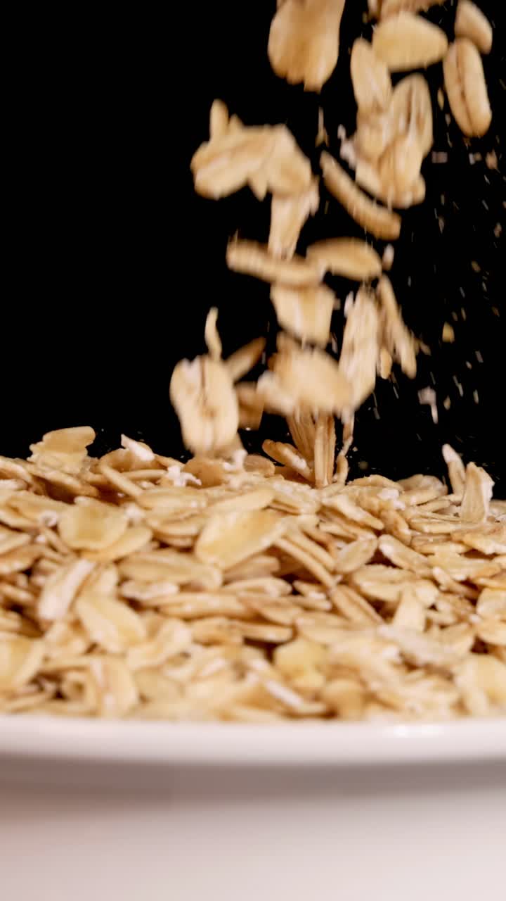 Chicchi d'avena cadono a cascata in una ciotola bianca su uno sfondo nero, ripresi al rallentatore con illuminazione drammatica.
