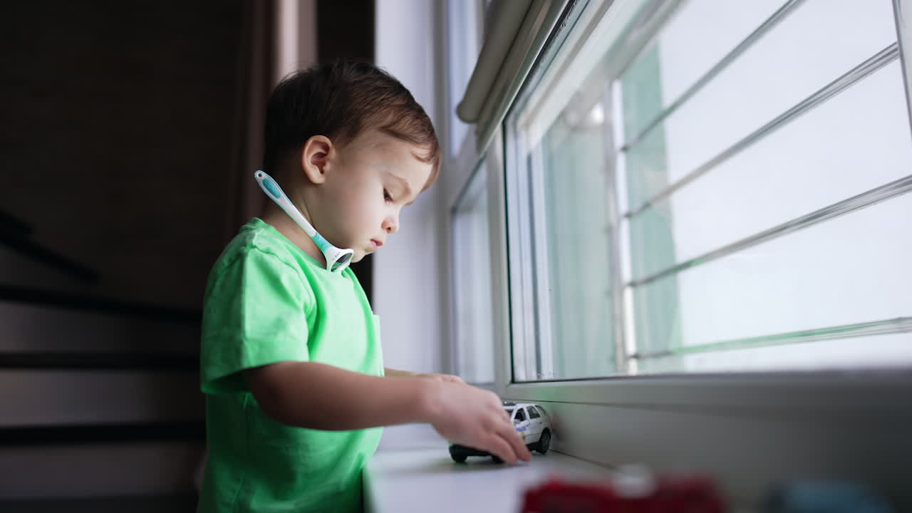 Little boy stands at the window. Cute kid is focused on toy police car pushing it by the window-sill.