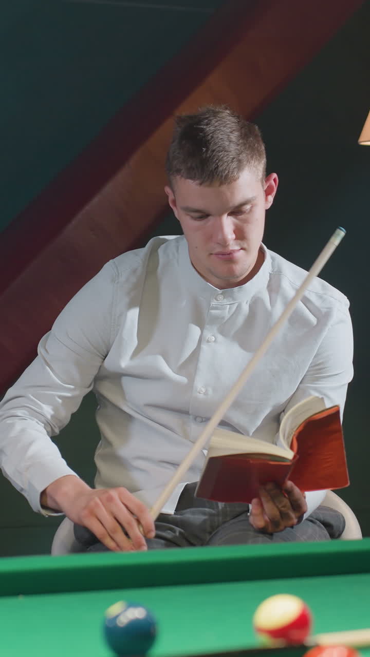 Mann i hvit skjorte sitter på en hvit stol og leser en rød bok mens han holder en køpinne nær et grønt biljardbord med spredte biljardkuler. Varm belysning fra en gulvlampe skaper en avslappet atmosfære.