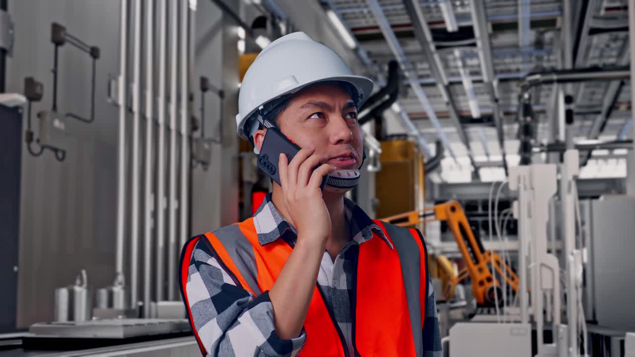 Primer plano de un ingeniero asiático con casco de seguridad hablando por teléfono inteligente mientras está de pie, línea de producción automatizada de baterías en una fábrica moderna