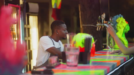 barman with beer poses to woman camera in pub. african american barkeeper waits while guest takes picture and looks at phone screen in night club