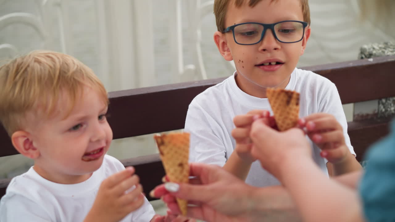 Two children sit on a bench with excitement as they reach out toward ice cream being handed to them, the younger child enthusiastically points, while the older sibling watches