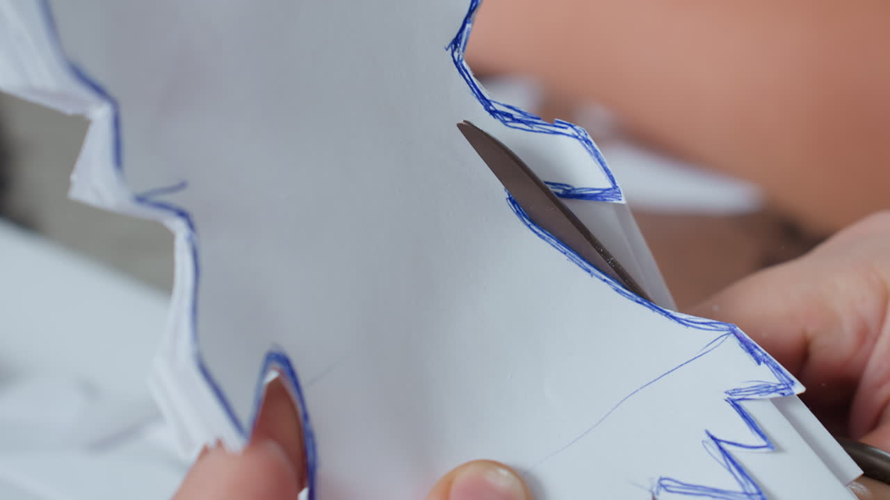 close hand view carefully using scissors cutting along blue outlined shapes on white paper while creating handmade craft project on table with scattered paper pieces during indoor creative session