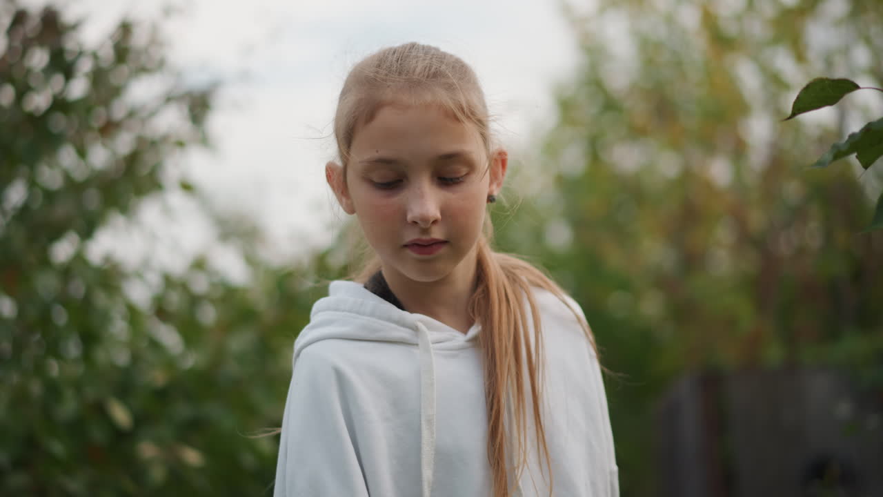 Jeune fille caucasienne dans un jardin, portant un sweat à capuche blanc, le regard baissé avec une expression douce, faible profondeur de champ, bokeh vert chaud, début de l'automne, lumière naturelle, portrait intime, sérénité émotionnelle, enfance.