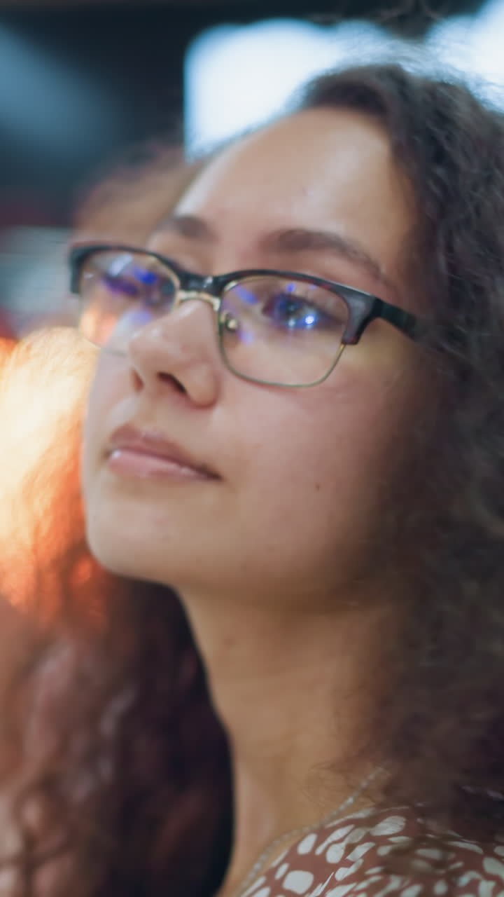 primer plano de una mujer con cabello rizado y gafas sentada en el centro comercial, mirando cálidamente a la distancia con una sonrisa suave, rodeada de luces bokeh suaves y borrosas en el fondo