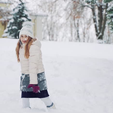 A beauty girl in a warm down jacket and hat holds a snowball in her hand and throws it forward on a cold weather in the winter in the hill near the house. Slow motion