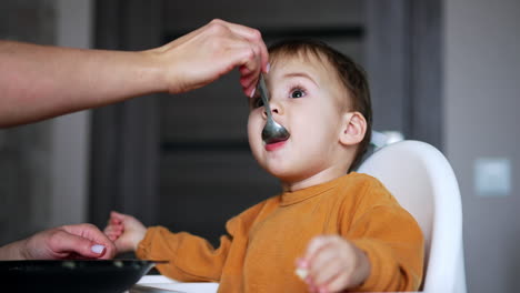 adorable face of a lovely baby eating porridge from spoon. feeding a kid at home. close up. blurred backdrop.