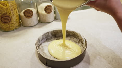 home cook slowly pours creamy cake mixture into round lined baking tray