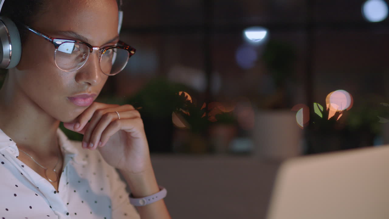 mujer de negocios de raza mixta que trabaja hasta tarde usando una computadora portátil escribiendo gerente de proyecto navegando en línea planeando la investigación fecha límite escuchando música en la oficina por la noche