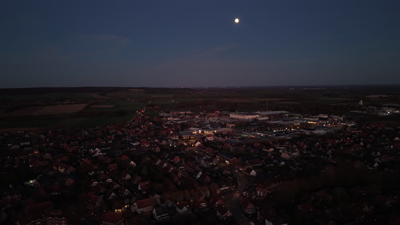 Luftaufnahme mit Drohne einer kleinen amerikanischen Stadt bei Nacht unter hellem Vollmond, mit Häusern, Straßen und entfernter Landschaft sichtbar am Abendhimmel. Weitwinkel. Panorama.