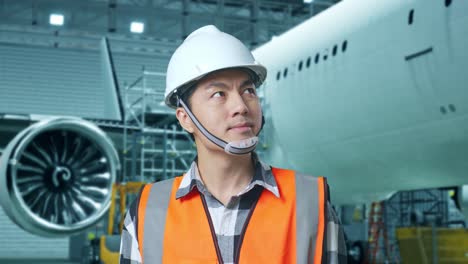 primer plano de un ingeniero asiático con casco de seguridad de pie con aviones en el hangar. mirando a su alrededor, comprobando