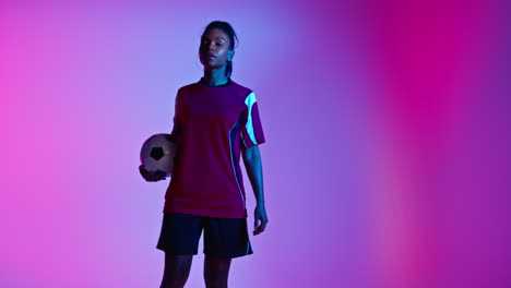 Studio-Portrait-Of-Female-Football-Or-Soccer-Player-Wearing-Team-Shirt-Holding-Ball-Under-Her-Arm-Shot-Low-Key-Against-Colourful-Mixed-Lighting-3