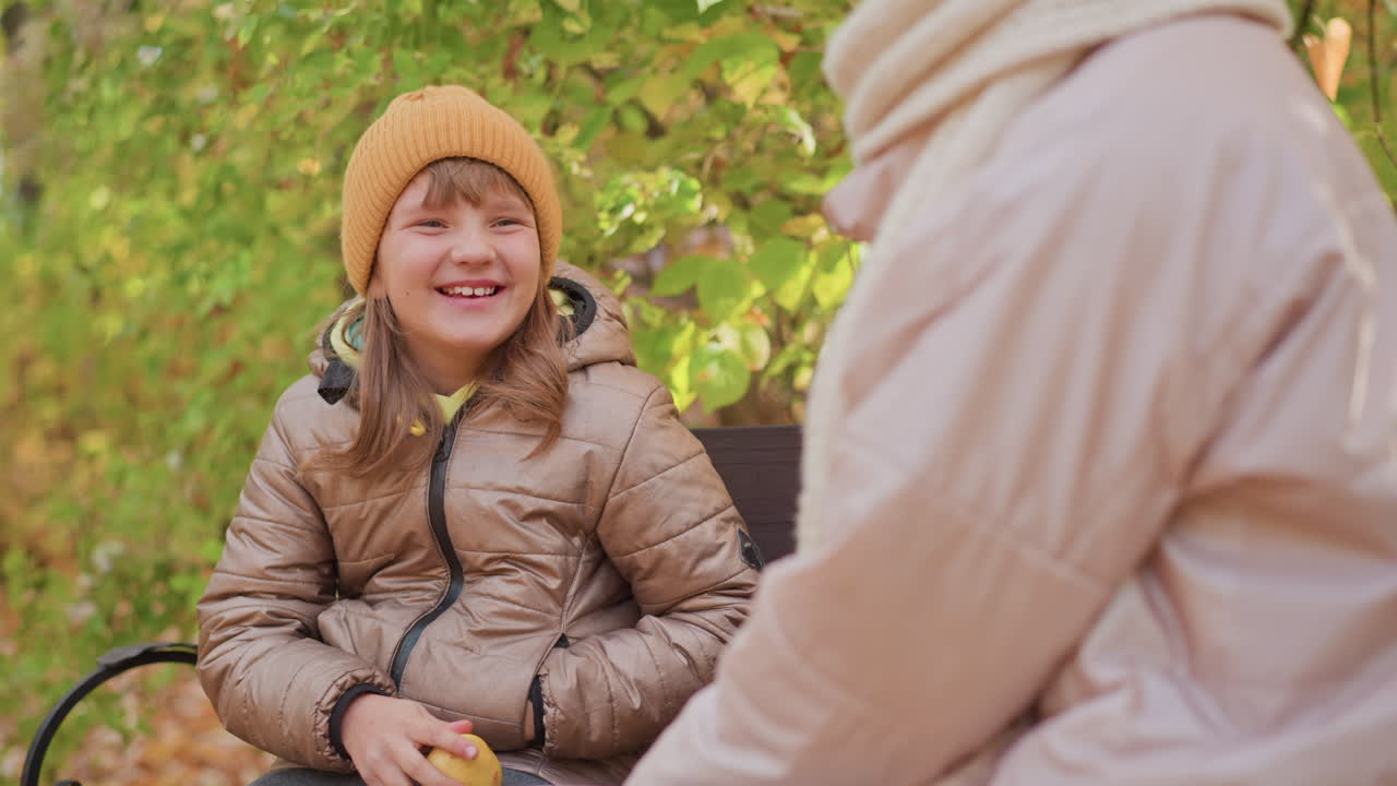 young girl in orange beanie holds apple with hands and smiles warmly at mother sitting nearby on park bench under golden autumn leaves sharing tender moment of parent child bonding outdoors