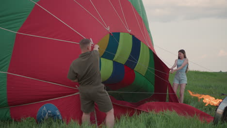 couple hold hot air balloon envelope open while burner flame heats fabric as wind inflates vibrant checker pattern during calm evening preflight setup in grassy field under pastel sky horizon