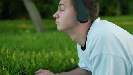 side view of man lying on grassy field wearing headphones, head slightly lifted, nodding rhythmically while enjoying music, surrounded by lush green grass and serene natural surroundings