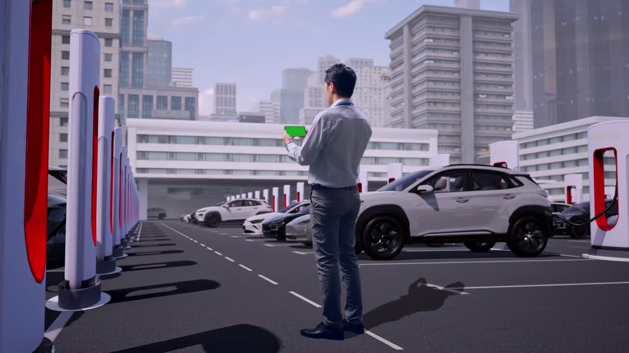 最新の都市型充電ステーションの工業施設で電気自動車が充電されている中、グリーンスクリーンのスマートフォンを手に立つアジア人男性の専門職作業員の全身の後ろ姿。