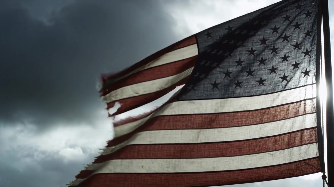 Una bandiera americana logora sventola, frustata da forti raffiche contro un cielo coperto, con i bordi strappati.