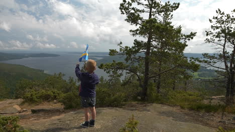 Young-Tourist-Waving-Swedish-Flag-on-Top-of-a-Mountain-Overlooking-High-Coast-of-Sweden