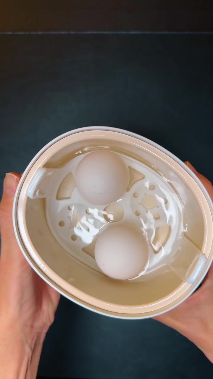 Pouring water into white electric egg cooker base. Top view of hand pouring water from glass into kitchen appliance base on dark background.