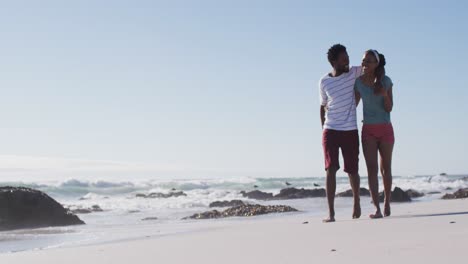 african american couple smiling, embracing and walking on the beach