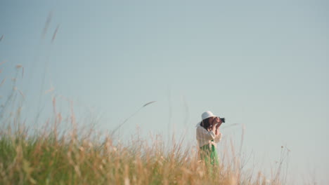 golden blades of grass sway gently in foreground while blurred woman with white hat walks through field in background capturing moment on camera under clear blue sky