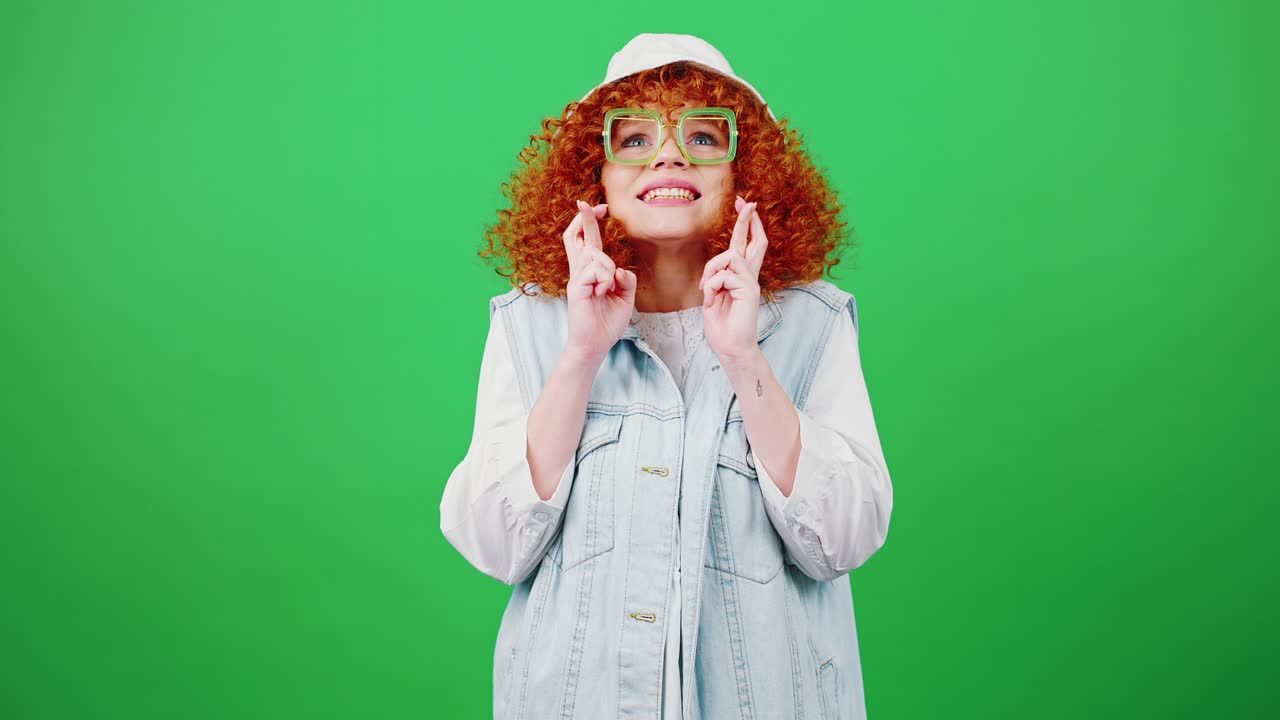 Young funny redhead curly woman in glasses and panama hat making cherished wish, crossing fingers, chroma key background