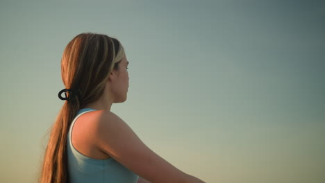 young woman in cyan signet standing outdoors with warm sunlight reflecting off her face, hair tied back, gazing into distance with focused expression