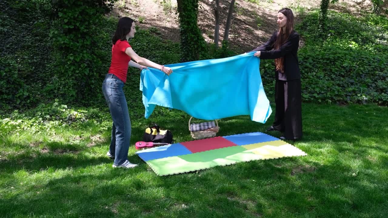 Jovens mulheres cobrem esteira com cobertor preparando local de piquenique no parque.
