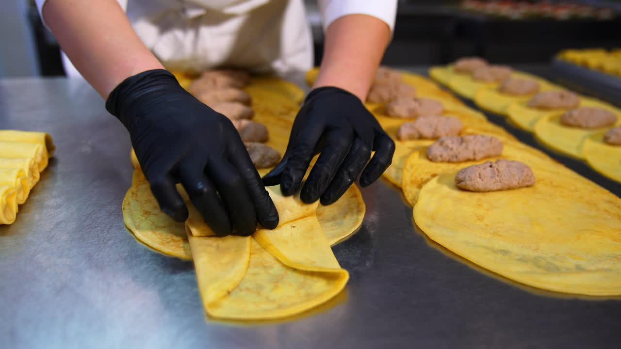 Hands of food factory worker roll the fried minced meat into a yellow crepe. Half-stuff food manufacturing handmade. Close up.