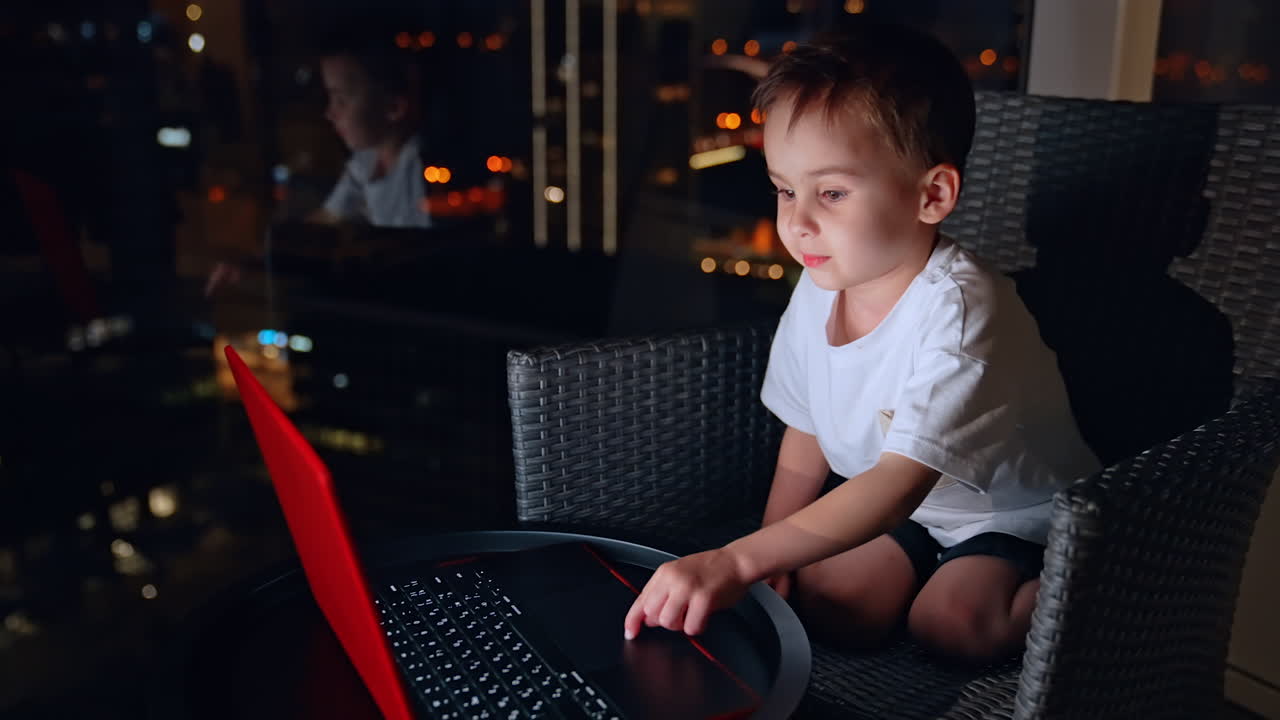 Small kid sits in the chair near the window in the dark room. Toddler boy is working on the laptop