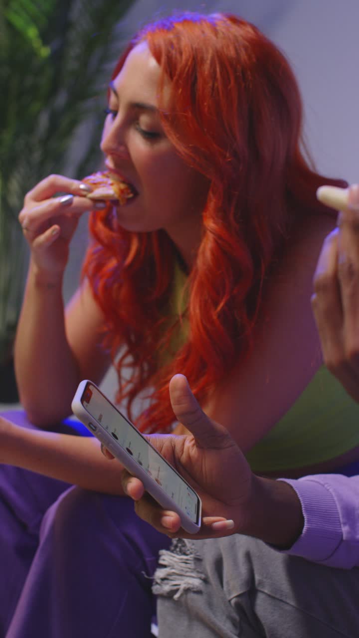 video vertical de un grupo de amigos de la generación z sentados en el sofá comiendo pizza para llevar a casa y usando teléfonos móviles 1