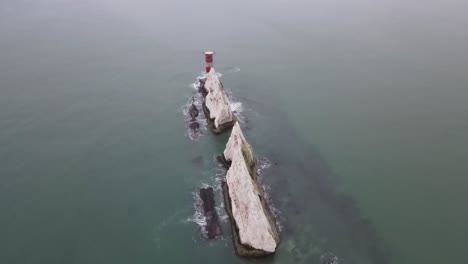 aerial drone flight in the isle of wight looking down on the red and white lighthouse at the needles with waves crashing against the white cliffs