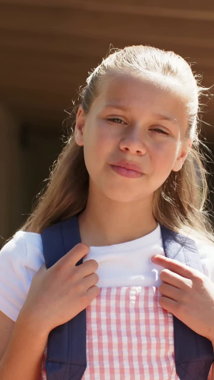 縦型動画：陽光に反応し、ギンガムチェックの服を着た少女がポーチでまばたきしながら微笑み、バックパックのストラップを調整している。