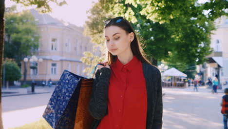 Eine-Attraktive-Frau-Mit-Einkaufstüten-Läuft-Durch-Die-Stadt,-Sie-Trägt-Ein-Rotes-Kleid-Und-Eine-Sonnenbrille