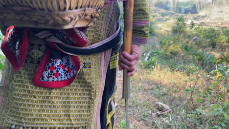 tracking shot of a black dao person walking through vegetation in traditional clothing, holding a harvesting tool.