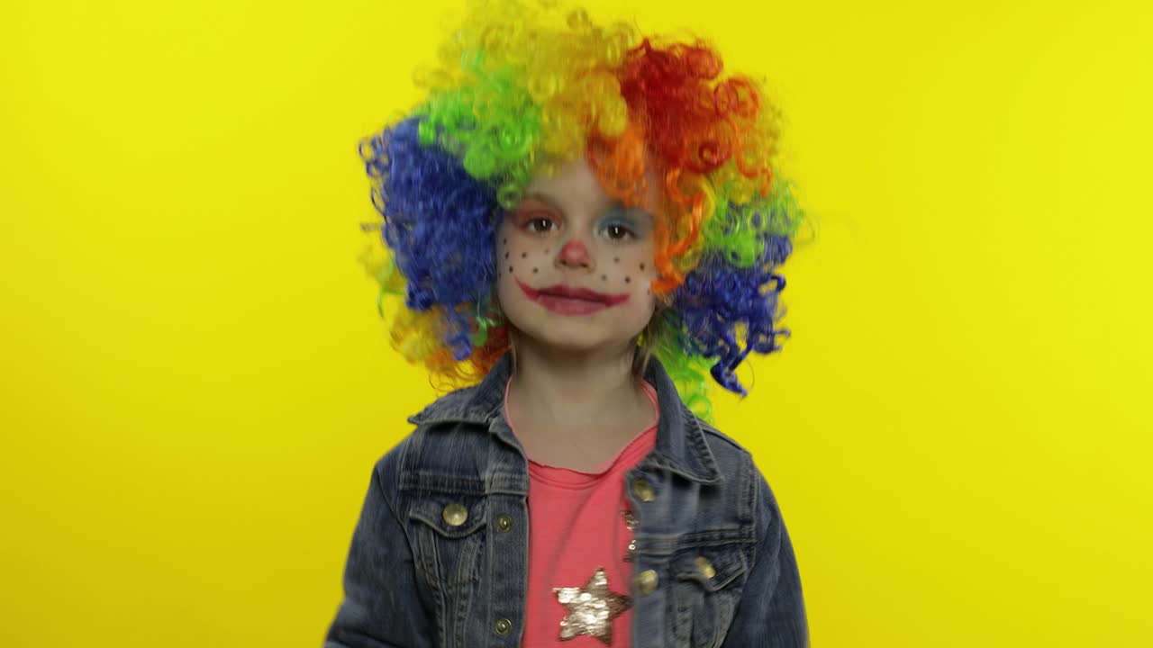 una pequeña niña payasa con una peluca colorida haciendo caras tontas, cantando, sonriendo, bailando. halloween