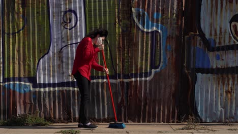 Woman-with-a-disposable-face-mask-and-red-cardigan-sweeps-the-street,-dirt-dust-in-the-air