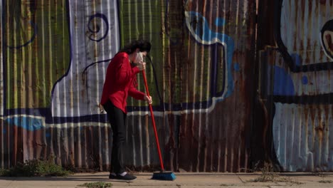 una mujer con una mascarilla desechable y una rebeca roja barre la calle, polvo de tierra en el aire
