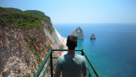 Male-walking-out-onto-a-catwalk-on-the-edge-of-a-cliff-over-looking-a-beautiful-ocean-landscape-with-clear-blue-water