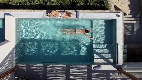 Brunette-woman-in-swimming-costume-lying-at-the-edge-of-the-pool-at-Cayo-Resort-in-Greece-Crete---man-gives-her-a-kiss-and-swims-in-the-pool---drone-flies-over-them-in-bird's-eye-view