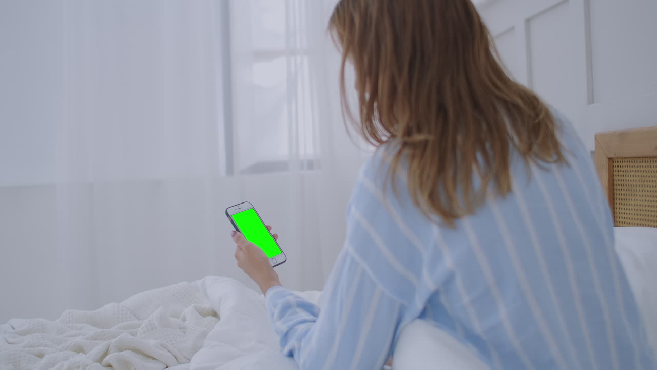 Young woman with cell phone on bed shoulder view. Technology addiction. Green screen phone. Young woman touching belly and using smartphone in bedroom