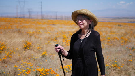 una anciana de cabello gris caminando en un campo de flores naranjas en una caminata con su bastón a cámara lenta